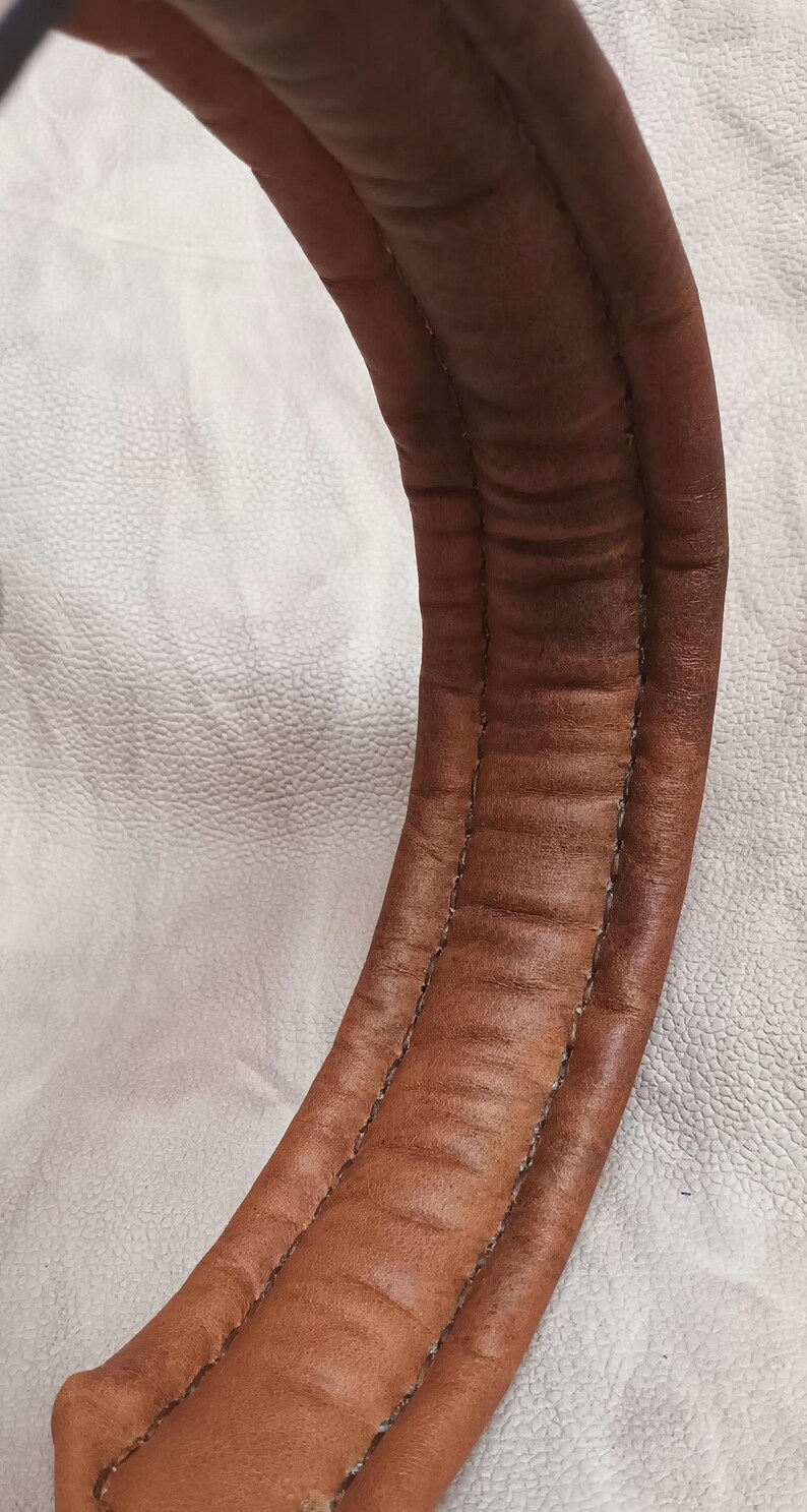 May include: Close-up of a curved, brown leather object with visible stitching. The leather has a textured appearance with darker areas, suggesting wear. The object is set against a white, textured background.