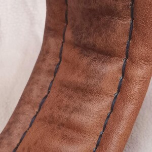 May include: Close-up of brown leather with a black stitched seam. The leather has a textured surface.