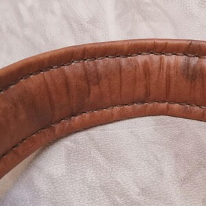 Puede incluir: Primer plano de un asa de cuero marr&oacute;n curvada con costuras visibles. El cuero tiene un aspecto desgastado con &aacute;reas m&aacute;s oscuras, lo que sugiere edad o uso. El asa es probablemente parte de una bolsa o art&iacute;culo similar.