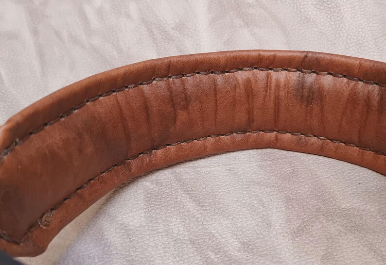 Puede incluir: Primer plano de un asa de cuero marr&oacute;n curvada con costuras visibles. El cuero tiene un aspecto desgastado con &aacute;reas m&aacute;s oscuras, lo que sugiere edad o uso. El asa es probablemente parte de una bolsa o art&iacute;culo similar.