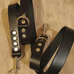 Könnte beinhalten: Zwei schwarze Lederhundeleinen mit silberner Beschläge. Eine Leine hat einen D-Ring und einen Karabinerhaken, die andere hat einen Karabinerhaken und einen Ring mit fünf silbernen Nieten.