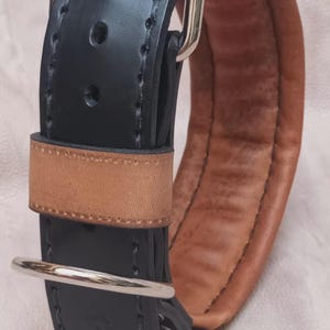 May include: A black leather dog collar with a brown leather lining and a silver buckle. The collar is stitched with white thread.