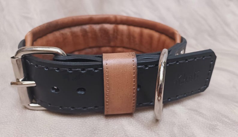 May include: A black leather dog collar with a brown leather lining and a silver buckle. The collar has a stitched design and the word "K9" is embossed on the leather.