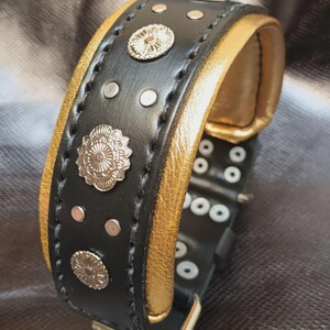 May include: Black leather dog collar with gold accents and silver metal embellishments. The collar has a buckle closure and multiple rows of stitching.