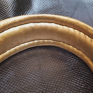 May include: Close-up of a gold-colored leather strap with a stitched seam. The strap is curved and has a smooth, polished finish.