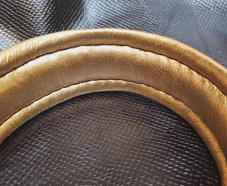 May include: Close-up of a gold-colored leather strap with a stitched seam. The strap is curved and has a smooth, polished finish.