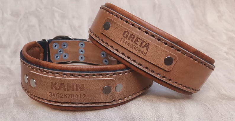 Puede incluir: Dos collares de perro de cuero marr&oacute;n. Uno est&aacute; abierto, el otro apilado encima. Cada uno tiene un nombre y un n&uacute;mero de tel&eacute;fono grabados en un parche rectangular. El collar abierto tiene "KAHN" y "3462670412". El otro tiene "GRETA" y "1144030848".