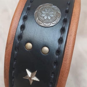 May include: Black leather dog collar with brown leather backing. The collar has silver star and circle accents with a decorative design.