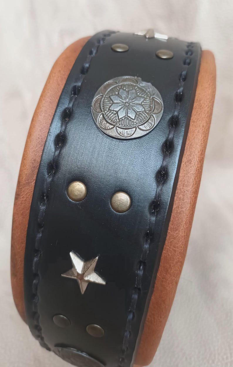 May include: Black leather dog collar with brown leather backing. The collar has silver star and circle accents with a decorative design.