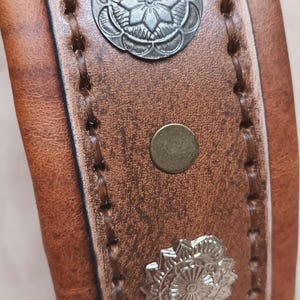 May include: Close-up of a brown leather dog collar with decorative silver floral conchos and brass rivets. The collar has stitched edges and a buckle. The leather has a distressed appearance.