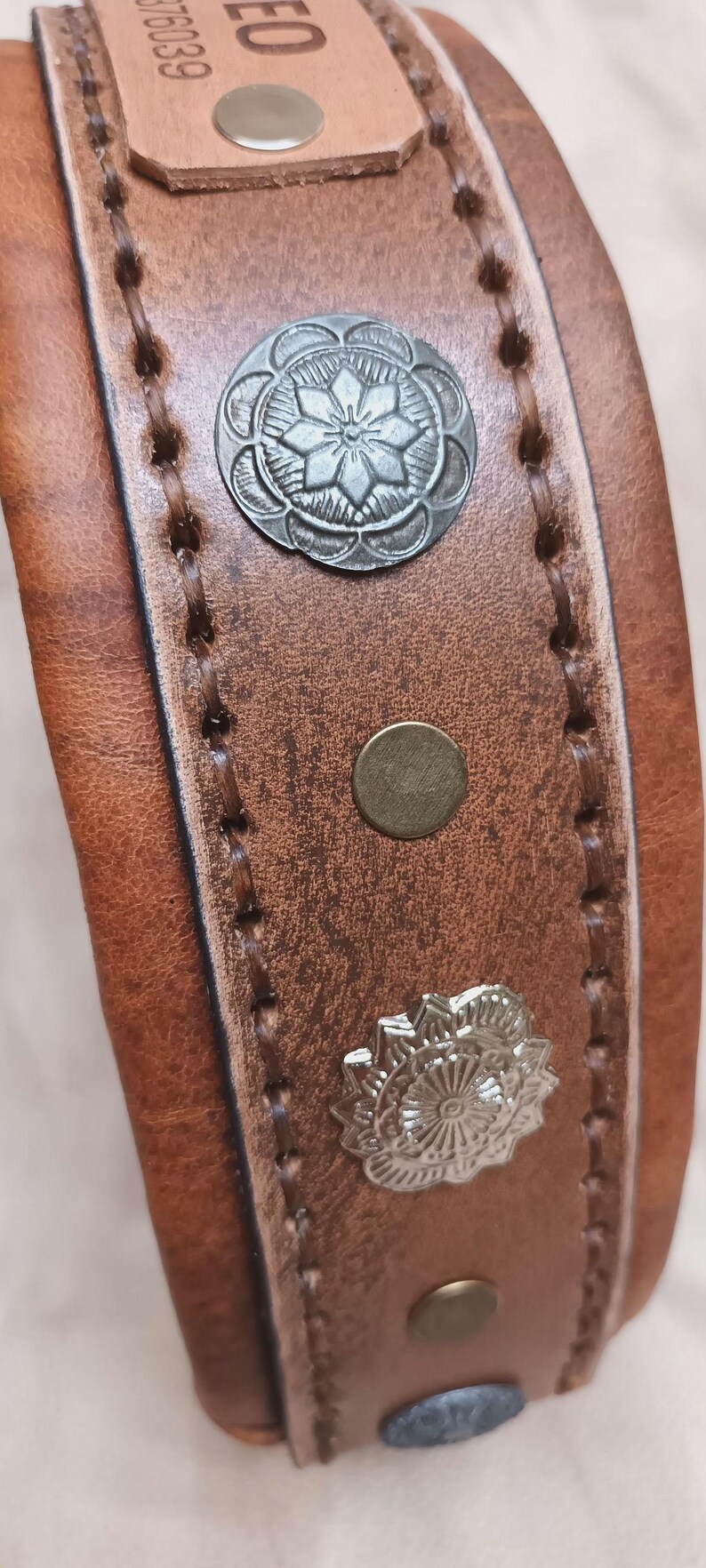 May include: Close-up of a brown leather dog collar with decorative silver floral conchos and brass rivets. The collar has stitched edges and a buckle. The leather has a distressed appearance.