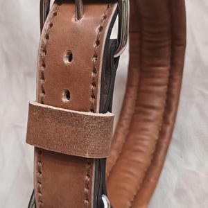 May include: A close-up of a brown leather dog collar with a padded interior. The collar has a silver-coloured buckle and a D-ring for leash attachment. The leather is stitched with a contrasting thread.