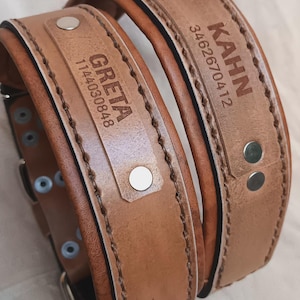 Puede incluir: Dos collares de perro de cuero marr&oacute;n claro con placas de identificaci&oacute;n. Uno dice "GRETA" con un n&uacute;mero, el otro "KAHN" con otro n&uacute;mero. Ambos collares tienen remaches y hebillas plateadas.