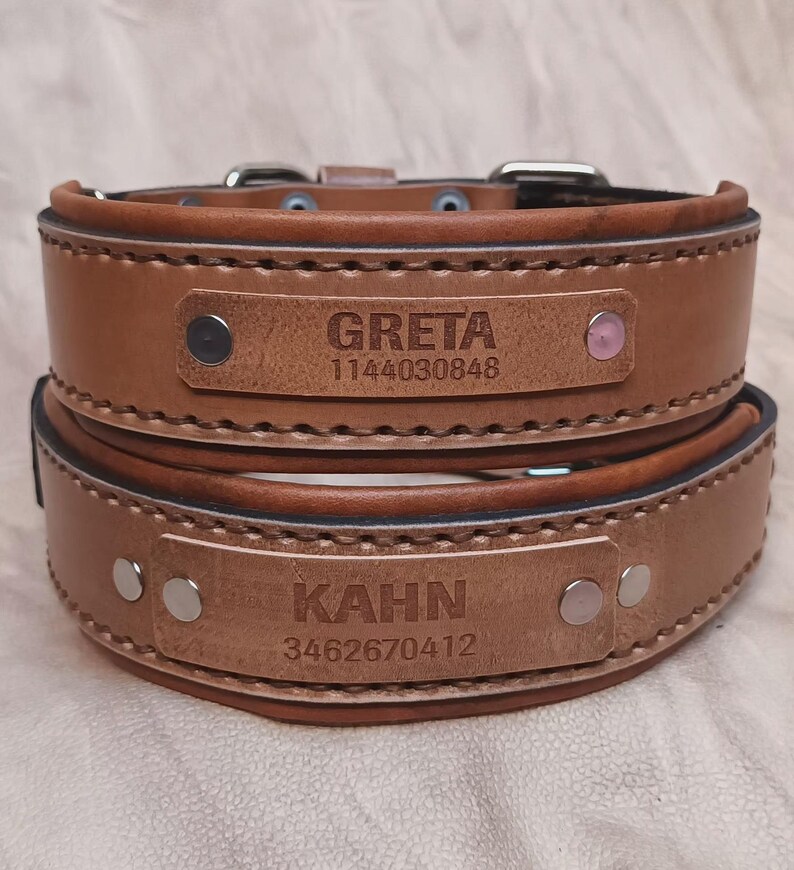 Puede incluir: Dos collares de perro de cuero marr&oacute;n apilados uno encima del otro. Cada collar tiene una placa con el nombre de un perro y un n&uacute;mero de tel&eacute;fono. El collar superior dice "GRETA" y el collar inferior dice "KAHN".