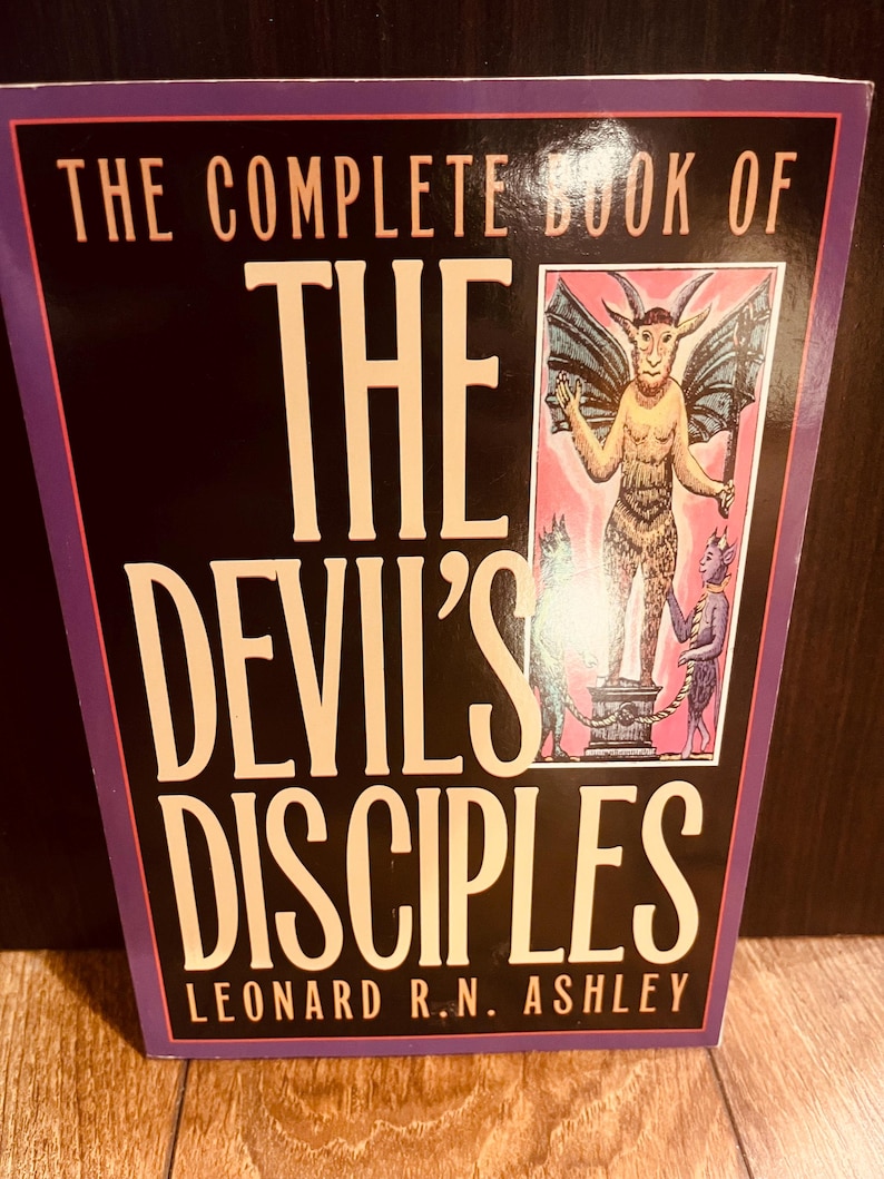 Pu&ograve; includere: Un libro intitolato "THE COMPLETE BOOK OF THE DEVIL'S DISCIPLES" di Leonard R.N. Ashley. La copertina presenta uno sfondo nero con testo bianco e un bordo viola. Un'illustrazione di una figura simile al diavolo &egrave; sul lato destro.