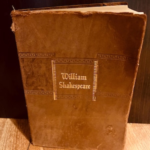 May include: A vintage, brown leather-bound book titled "William Shakespeare" in gold lettering. The book has a worn appearance, with visible wear and tear on the cover and edges. The cover features a decorative border.