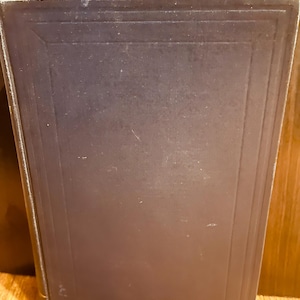 May include: A vintage book with a dark blue cover. The cover has a rectangular design with embossed lines. The book's spine is visible on the left side, with gold-colored text. The book is slightly angled.
