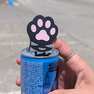 Op de afbeelding: Een zwarte en roze kattenpoot-bliktopper is in een blauw energiedrankblikje gestoken. Op het blikje staat "SUGAR ENERGY". Het blikje wordt door een persoon vastgehouden.