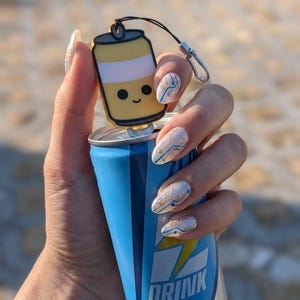 Pode incluir: Uma lata de bebida energética azul e prateada com a palavra "DRINK" em branco. Um pingente de lata de desenho animado amarelo e branco com um rosto sorridente está preso à lata. A lata é segurada em uma mão com unhas decoradas.