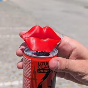 Pode incluir: Um abridor de latas vermelho em forma de lábios, impresso em 3D, está em cima de uma lata de bebida energética vermelha e preta. A lata apresenta o texto "URB CLASS ENERGY" e um gráfico de arranha-céus. O abridor é segurado por uma mão.