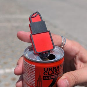 Pode incluir: Um abridor de latas em forma de batom vermelho e preto é colocado em cima de uma lata vermelha de Urban Classic Energy Drink. O abridor de latas é feito de plástico e tem um design detalhado. A lata é segurada por uma pessoa.