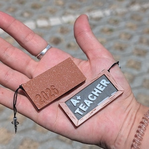 Peut inclure: Deux petites étiquettes rectangulaires marron sont tenues dans une main. L'une porte l'année en relief "2026". L'autre étiquette a la forme d'un tableau noir avec les mots "A+ TEACHER" en lettres blanches. Les deux étiquettes ont une ficelle noire attachée.