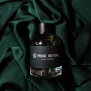Può includere: Un flacone di profumo in vetro trasparente con un tappo nero e un'etichetta. L'etichetta riporta la scritta "PRIME REVERIE LUNA CUBANA". Il flacone è appoggiato su uno sfondo di tessuto verde.