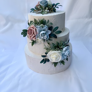 May include: A three-tiered white wedding cake decorated with blush pink, white, and blue flowers and greenery.