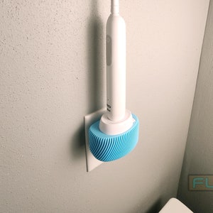 May include: A white electric toothbrush with a blue silicone charging base mounted on a white wall outlet.