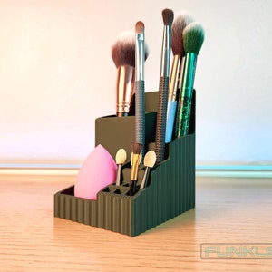 May include: A dark green, tiered makeup brush organizer with vertical ribbed sides. It holds various makeup brushes with metallic and glitter handles, along with a pink teardrop-shaped makeup sponge and three small foam-tipped applicators.