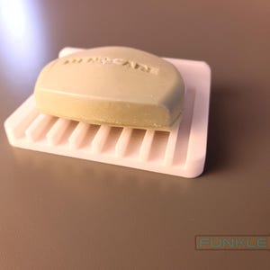 May include: A white soap dish with a raised grid pattern and a green bar of soap on top. The soap has the words "All Natural" printed on it.