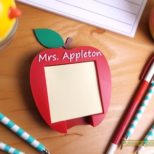 May include: A red apple-shaped memo holder with the name "Mrs. Appleton" in white. The holder has a blank, cream-colored notepad. Pens with red and teal stripes are nearby, along with a yellow bird-shaped object.