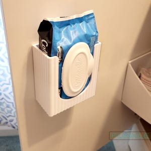 May include: White plastic dispenser for a package of blue and black Clorox wipes. The dispenser is mounted on a white wall.
