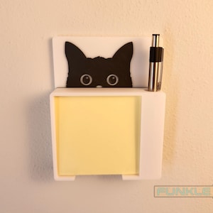 May include: White wall mounted note holder with a black cat peeking out from the top. The holder has a yellow sticky note pad and a silver pen.