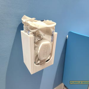 May include: A white, 3D-printed wall-mounted wipe dispenser. The dispenser holds a package of wipes, with the lid visible. The dispenser is mounted on a light blue wall. A blue crib is in the background.