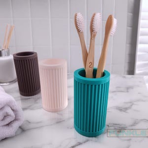 May include: Three bamboo toothbrushes with white bristles stand in a teal blue silicone toothbrush holder. Two additional holders are visible in the background, one brown and one pink.