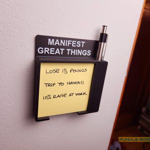 May include: A black wall-mounted organizer with the text "MANIFEST GREAT THINGS." It holds a yellow sticky note with handwritten goals: "LOSE 15 POUNDS," "TRIP TO HAWAII," and "10% RAISE AT WORK." A pen is in the holder.