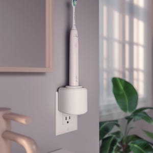 May include: A white electric toothbrush is mounted on a white charging base plugged into a wall outlet. The toothbrush is light pink with a green and white brush head. The scene is set in a bathroom with a mirror and a plant visible.