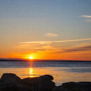 May include: A scenic sunset over a calm body of water. The sky transitions from blue to orange and yellow, with the sun low on the horizon. Dark rocks are in the foreground, silhouetted against the water.