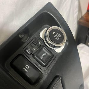 May include: Black plastic car interior panel with a silver engine start stop button, a mirror control button, and a headlight control button. The button says "Engine Start Stop".