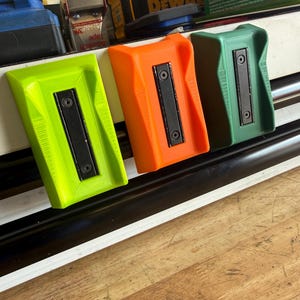 May include: Three magnetic tool holders in neon green, bright orange, and dark green. Each holder features a rectangular black magnetic strip secured by two screws, with a textured, angled plastic body designed for mounting on metal surfaces.