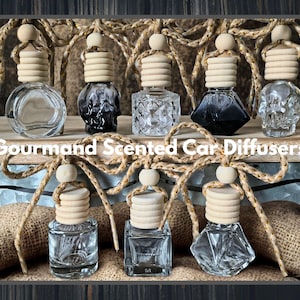 May include: An assortment of Gourmand Scented Car Diffusers in various clear glass bottles with wooden tops. Some bottles have black or skull-shaped designs. The bottles are suspended by rope and wood beads, with the text "Gourmand Scented Car Diffusers".