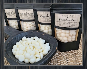 May include: An assortment of white wax melts in a black wooden scoop and black pouches. The pouches have labels with text like "Aged Barn", "Fall Farmhouse", "Cozy Flan", and "Birchwood Pine". The Birchwood Pine label lists ingredients.
