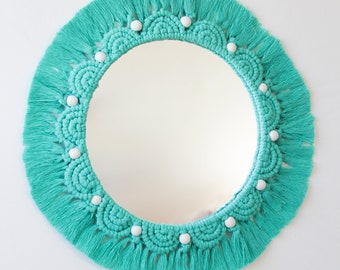 May include: A turquoise macrame mirror with white beads around the edge. The mirror has a fringe border.