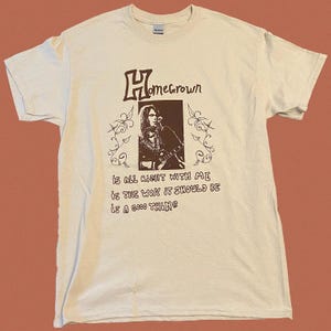 Könnte beinhalten: Ein beiges T-Shirt mit einem braunen Grafikdruck eines Musikers und dem Text "Homegrown" über dem Bild. Der Text unter dem Bild lautet "is all right with me is the way it should be is a good thing".