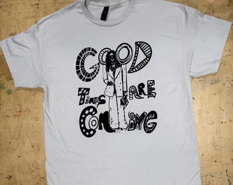 May include: A light gray short-sleeve t-shirt featuring a black graphic print of a person in a suit with the text "GOOD TIMES ARE COMING" in a stylized, bold font. The design includes decorative patterns within the letters and around the figure.