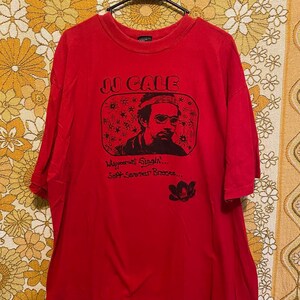 Könnte beinhalten: Rotes T-Shirt mit einem Schwarzweißbild des Musikers J.J. Cale. Das Bild ist in Form eines Fernsehbildschirms mit dem Text "Whipperwill Singin' Soft Summer Breeze..." darunter.