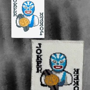 May include: A white rectangular playing card and a matching square crocheted textile piece, both featuring a pixelated graphic of a blue-masked wrestler holding a gold championship belt with the word JOKER printed vertically on both sides.
