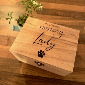 May include: A light-coloured wooden box with the words "IN LOVING memory Lady" in black script. A paw print is below the word "Lady". The box has a small metal clasp. A small plant in a blue watering can is in the background.