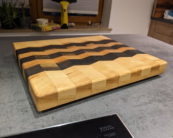 May include: A large, rectangular chopping board made of wood with a chevron pattern. The board is made of light wood, dark wood, and black wood.